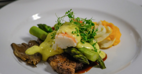 A gourmet dish featuring grilled meat, asparagus, mashed potatoes, and a creamy sauce, garnished with microgreens.