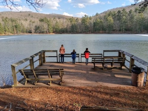 A woman and two children stand on a dock overlooking a frozen lake surrounded by trees and mountains.