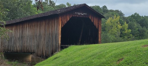 A rustic wooden covered bridge surrounded by green trees and grass under a cloudy sky.