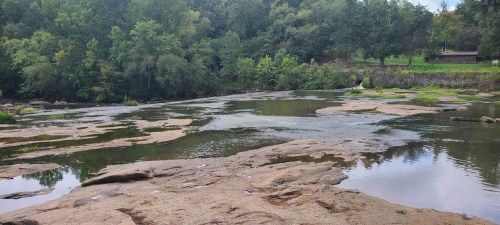 A serene river scene with rocky banks, surrounded by lush green trees and a calm water surface reflecting the landscape.