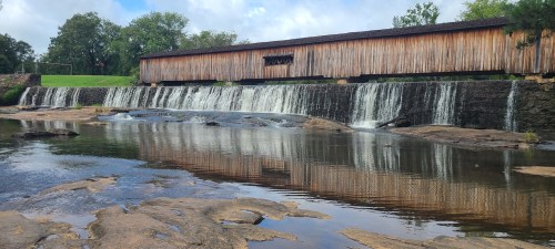 A wooden covered bridge spans a waterfall, reflecting in the calm water below, surrounded by greenery and blue skies.