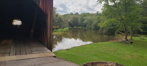 View from a covered bridge overlooking a calm river and green lawn with trees under a partly cloudy sky.