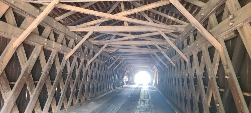 A wooden covered bridge interior with a crisscross pattern, leading to a bright light at the end.