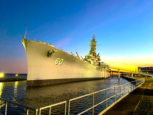 A battleship docked at sunset, with a clear sky and water reflecting the warm colors of the evening.