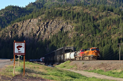 A train travels along a track beside a mountain, with a sign indicating a railroad crossing and fishing access nearby.