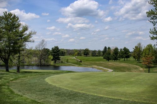 A scenic golf course with a green fairway, a pond, and trees under a partly cloudy sky.