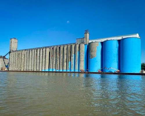 A large grain silo with blue-painted sections, standing by a calm body of water under a clear blue sky.