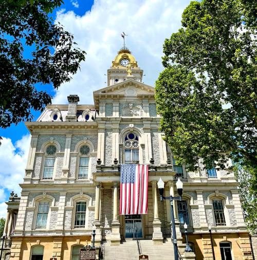Historic building with a gold dome, large American flag, and lush green trees under a bright blue sky.