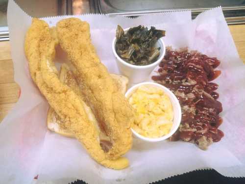 A plate featuring two fried fish fillets, collard greens, macaroni and cheese, and a serving of barbecue meat.