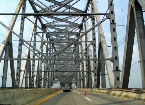View from inside a metal bridge, showcasing its structural beams and a car driving on the roadway below.