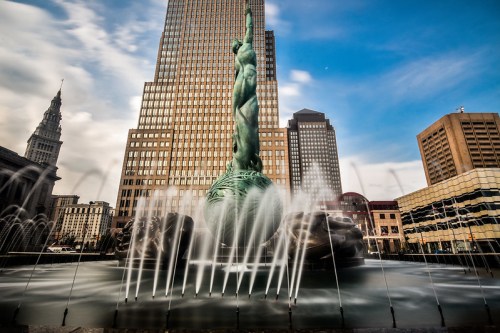 A statue resembling the Statue of Liberty stands in a fountain, surrounded by tall buildings under a blue sky.