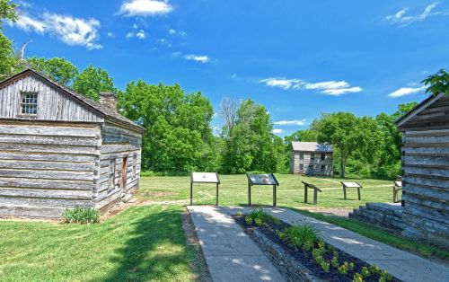 Two rustic log cabins with informational signs in a green landscape under a bright blue sky.