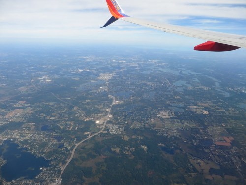 Aerial view of a landscape with lakes, roads, and towns, seen from an airplane wing against a blue sky.