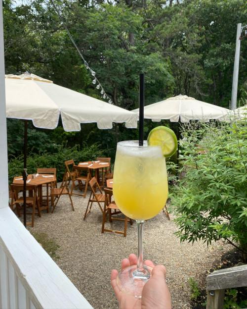A hand holds a yellow cocktail with a lime slice, overlooking a garden patio with tables and umbrellas.