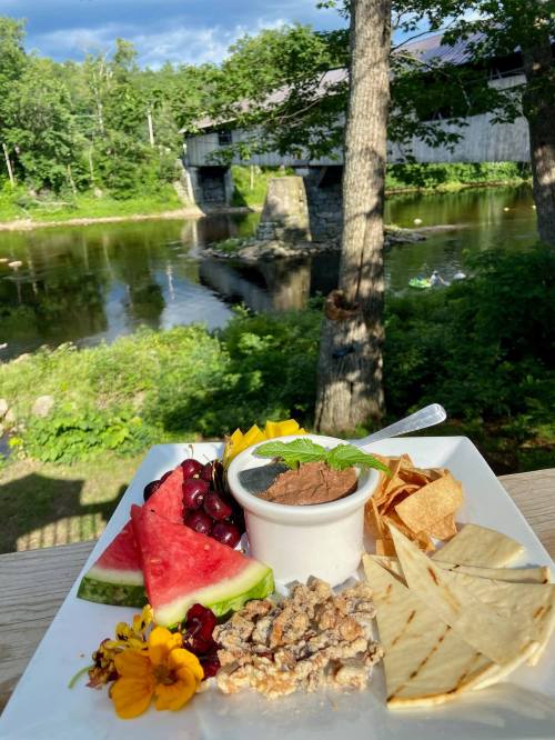 A plate of dessert with fruit, nuts, and chips, set against a scenic river and bridge backdrop.