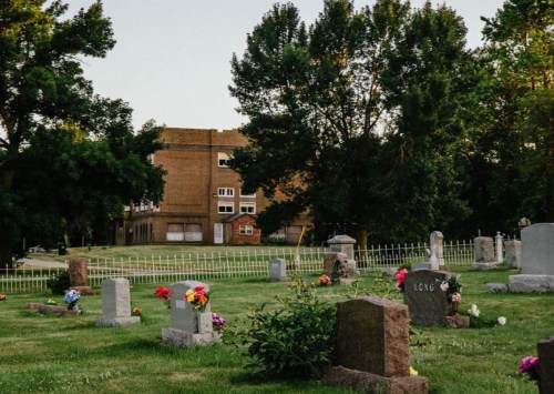 A serene cemetery with gravestones and colorful flowers, set against a backdrop of trees and a brick building.