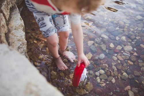 A child wades in shallow water, holding a red toy while exploring the rocky shoreline.