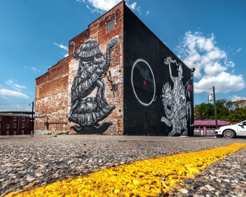 A vibrant mural on a brick building features large, intricate black and white characters against a blue sky.