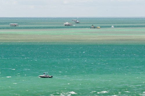 A small boat floats on turquoise waters, with distant structures visible on the horizon under a cloudy sky.