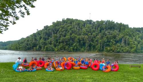 A group of people sitting on colorful inner tubes by a river, with a lush green hillside in the background.
