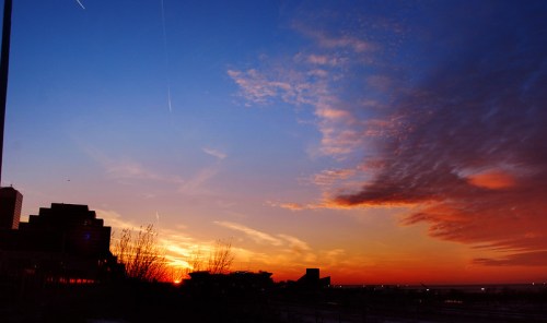A vibrant sunset with orange and blue hues, silhouetted buildings, and wispy clouds against a clear sky.