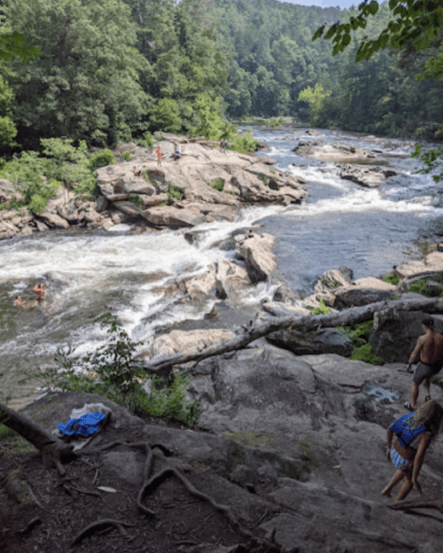 A scenic river with rocky banks, people swimming and exploring, surrounded by lush green trees and a clear blue sky.