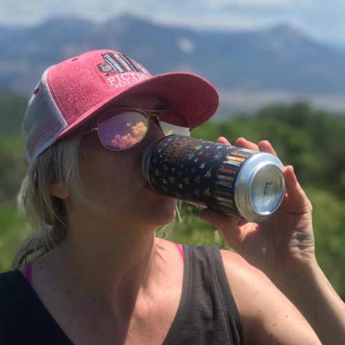 A person wearing a pink cap drinks from a colorful can, with mountains in the background on a sunny day.