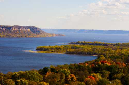 A scenic view of a river surrounded by lush greenery and autumn-colored trees under a clear blue sky.