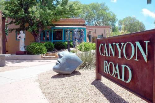 A sign reading "Canyon Road" in front of a building with sculptures and greenery in a sunny outdoor setting.