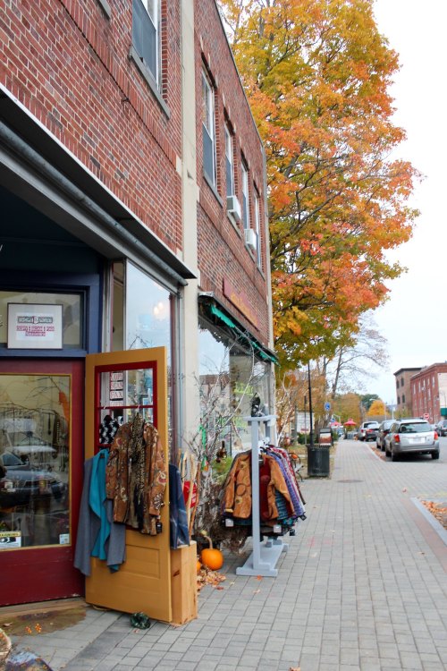 A quaint street scene featuring a brick building, colorful fall foliage, and a shop displaying various clothing items.
