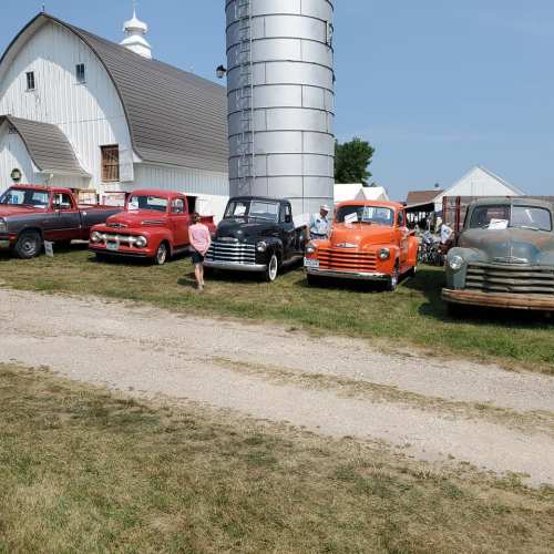 A row of vintage trucks parked beside a barn and silo on a sunny day, with people admiring the vehicles.