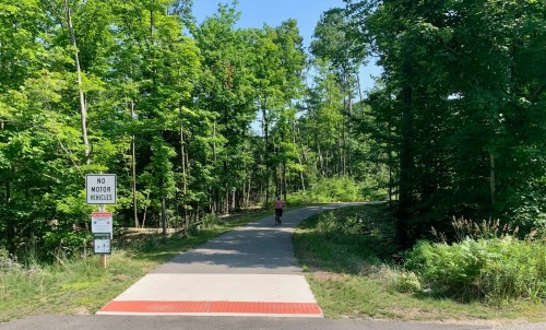 A paved path through a green forest, with a sign indicating no motor vehicles and a person walking in the distance.