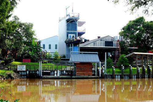 A riverside building with a tower, surrounded by greenery and a calm waterway, featuring docks and a modern design.