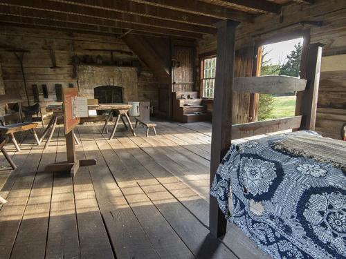 Interior of a rustic wooden room featuring a bed, table, and fireplace, with sunlight streaming through the windows.