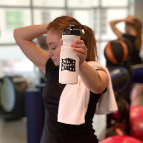 A person in a gym holds a water bottle with "Cherry Valley Hotel" printed on it, wearing a towel around their neck.