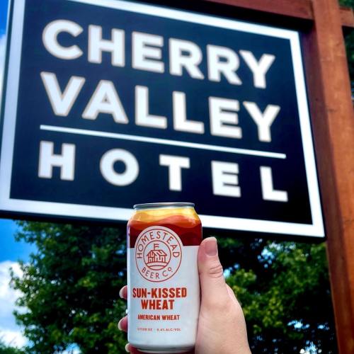 A hand holds a can of Homestead Beer Co. Cherry Kissed Wheat in front of the Cherry Valley Hotel sign.