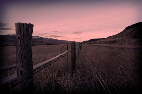 A desolate landscape at dusk, featuring a barbed wire fence, grassy fields, and distant mountains under a pink sky.