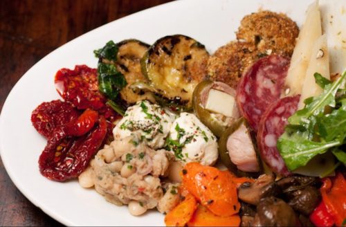 A colorful platter featuring grilled vegetables, cured meats, cheeses, olives, and various dips.