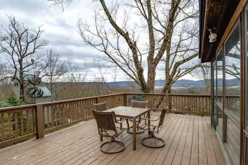 A wooden deck with a table and chairs overlooks a scenic view of trees and mountains under a cloudy sky.