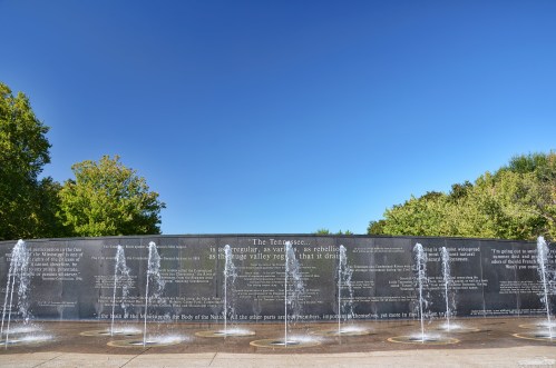 A stone wall with water fountains, featuring quotes and surrounded by trees under a clear blue sky.