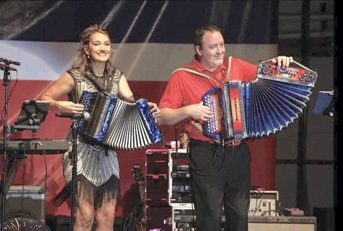 Two musicians on stage play colorful accordions, smiling and engaging with the audience. A band setup is visible behind them.