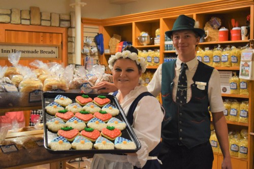 A woman in traditional attire holds a tray of decorated cookies, while a man in a vest smiles beside her in a bakery.