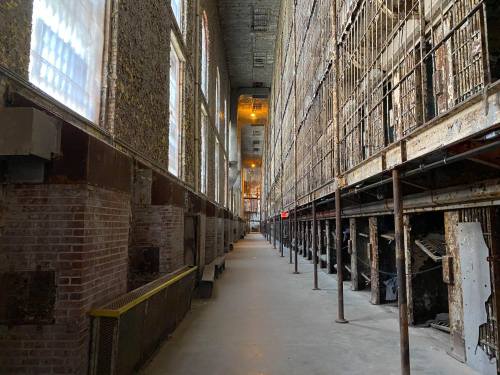 A long, dimly lit corridor in an old prison, featuring tall walls and rows of barred cells on either side.