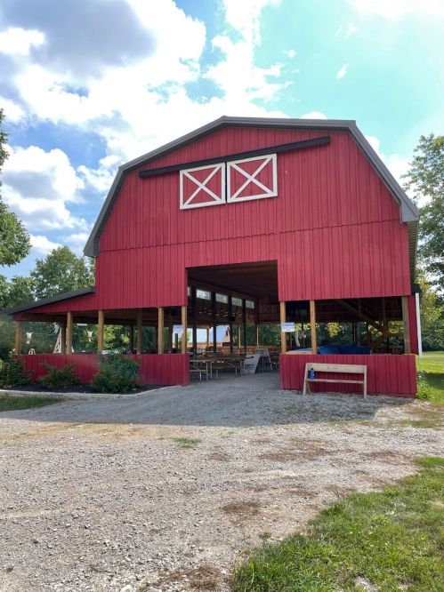 A red barn with a large open front, surrounded by grass and trees, under a partly cloudy sky.