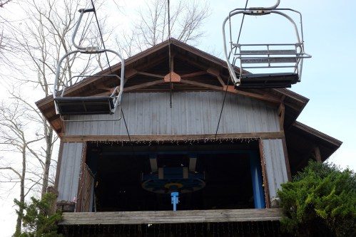 A wooden building with a sloped roof, featuring two empty ski lift chairs hanging above.