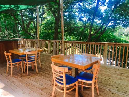 A wooden deck with two tables and chairs, surrounded by lush green trees and sunlight filtering through the leaves.
