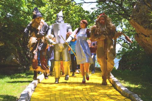 A group of people dressed as characters from "The Wizard of Oz" walking along a yellow brick road in a lush green setting.