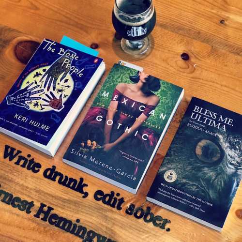 A wooden table with three books and a glass of dark beer. The titles include "The Bone People," "Mexican Gothic," and "Bless Me, Ultima."
