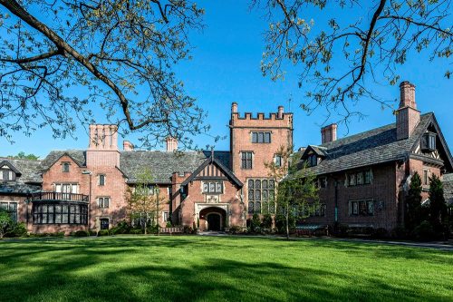 A large, historic brick mansion with towers, surrounded by green grass and trees under a clear blue sky.