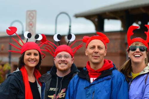 Four people wearing red hats with crab designs and playful expressions, posing together outdoors.
